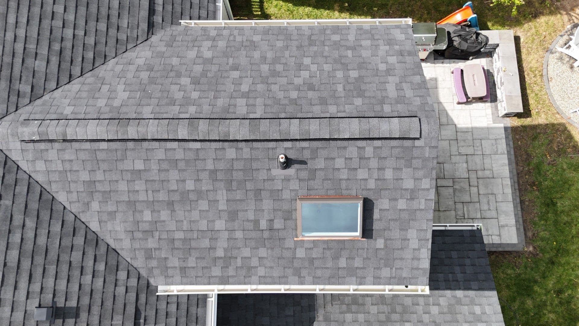 Aerial view of a residential roof with gray asphalt shingles, skylights, roof vents, and construction equipment and materials staged on the adjacent deck