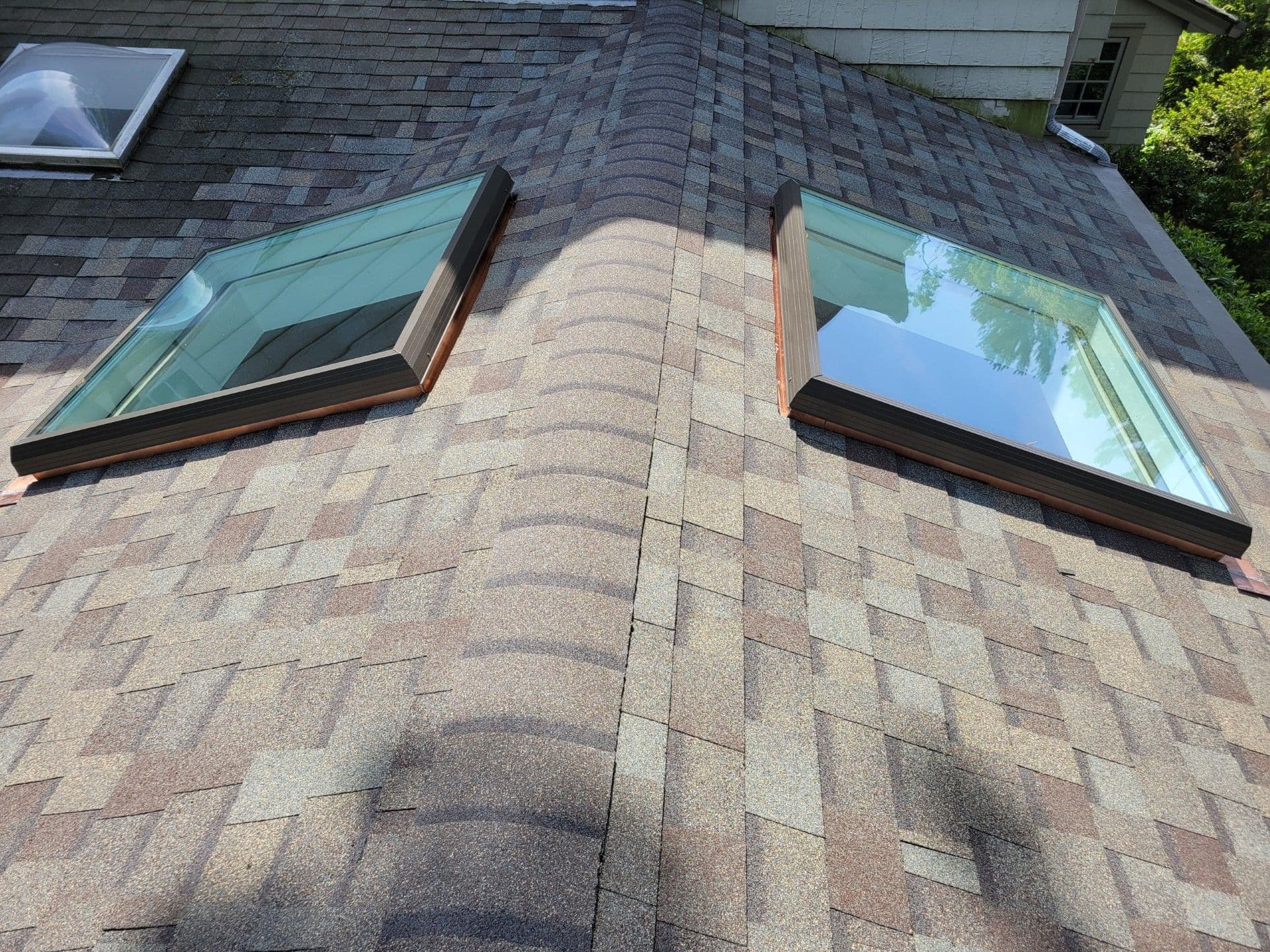 Roof with two skylights featuring teal frames and dark trim, positioned on asphalt shingles with a ridge line running between them